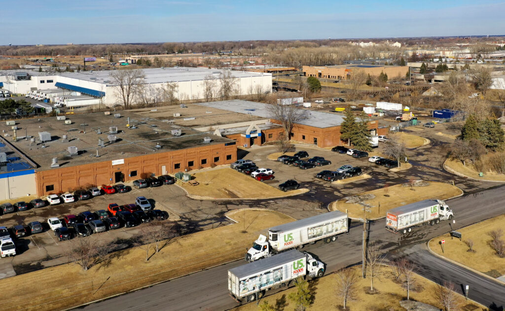 Knowledge Computers Drone picture of the Knowledge Computers commercial space from 200 feet in the air. In the foreground, three semi-trucks. The building is brick and there is a large parking lot in front with numerous cars.