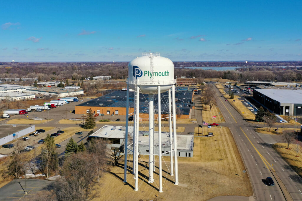Plymouth water tower. A drone shot of the Plymouth water tower. To the right of the water tower a road that stretches off to the distance. Blow and behind the water tower are commercial buildings.