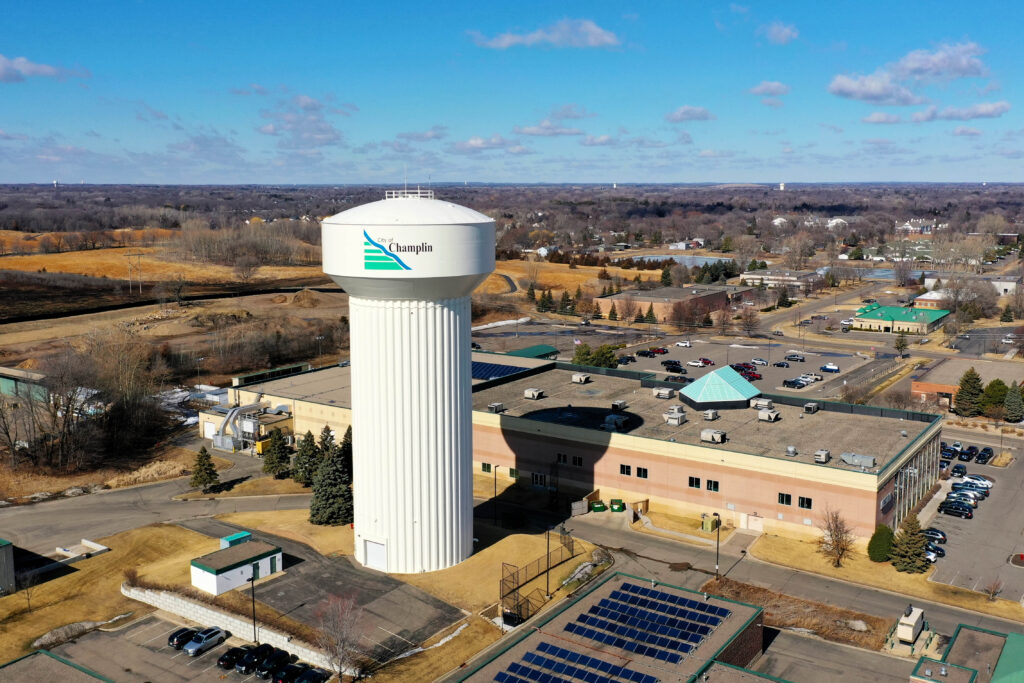 City of Champlin Water tower. City of Champlin Water tower shot by a drone, looking nearly eye level with the water tower with industrial buildings around it. In the distance, trees and blue skies with light clouds.